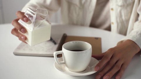 Joyful relaxed woman adds vegetable milk to coffee, Hand of a white girl pours milk from a milk jug into a cup with black hot coffee, a woman in a shirt sits at the table for breakfast
