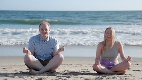 Beautiful couple enjoying healthy lifestyle during outdoor waterfront meditation. Cheerful man and woman sitting on sandy beach in front of calm ocean. High quality 4k footage