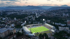 Cityscape featuring a stadium with green grass soccer field (football pitch) illuminated by floodlights at night, in the dark city of Podgorica, Montenegro, under colorful sunset sky in the evening. - Powered by Shutterstock - Get 15% off with code: PIKWIZARD15