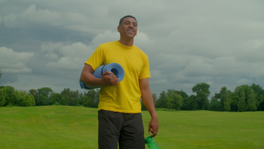 Positive handsome sporty fit African American man with yoga mat and sports water bottle in hands walking along green field. going for intense outdoor fitness workout in public park. | Shutterstock HD Video #1091631527