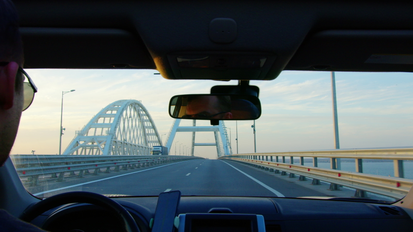 Man drives car on modern bridge surrounded by street lamps. View from inside automobile. Auto moving along road above river or bay. Transportation, travel, goods delivery, voyage, trip. Cloudy day Royalty-Free Stock Footage #1091634555