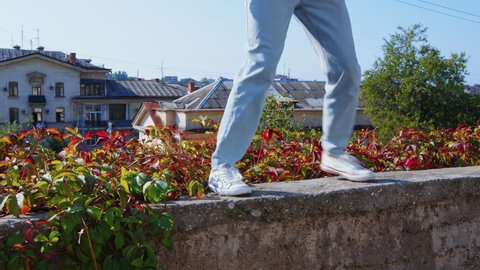Girl in jeans performing modern dances on retaining wall near flowerbed. Cozy town with small houses, roofs. Female dancer dancing hip hop, shuffle, contemporary. Woman rhythmically moving at street