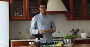 Happy handsome cook blogger man recording video on smartphone in kitchen, giving workshop, preparing salad, showing fresh vegetable ingredients at camera on tripod stand. Eating, marketing concept