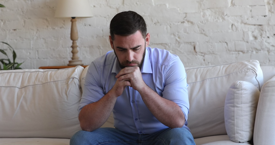 Stressed concerned millennial man thinking over bad news, problem solving, sitting on couch at home, touching, rubbing face, leaning chin on hands, feeling depressed, frustrated, coping with crisis | Shutterstock HD Video #1091639031