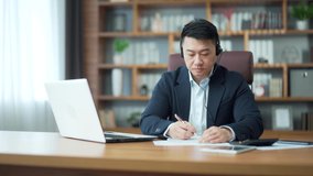 Asian employee studying online virtual tutor using laptop in office. distant worker wears headphones video calling makes notes, elearning with teacher, attending remote conference meeting training