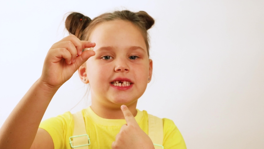 Little funny girl wide smile and point finger at gap in mouth from fallen tooth closeup, white background. Hold and show white milk tooth in hand. Concept of healthy teeth and beautiful smile. | Shutterstock HD Video #1091647525