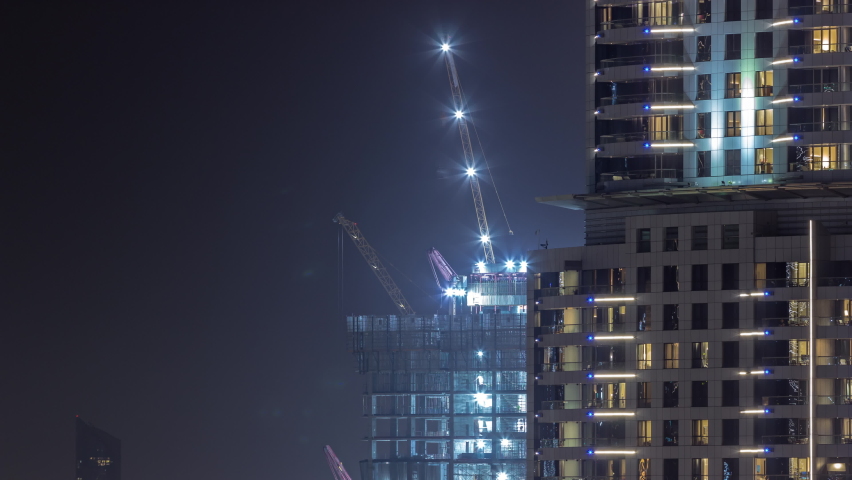 Tall buildings under construction and cranes in downtown night timelapse. Work progress at construction site of new towers behing skyscraper with glowing windows