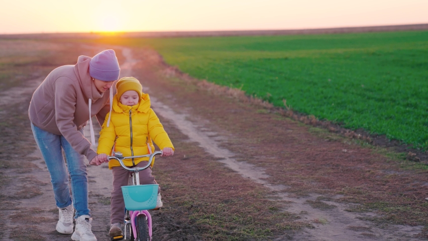 Family weekend, Mom teaches kid, daughter to ride bicycle in spring along country road. Happy family. Kid, girl learns to ride bike with her mother in park on green meadow at sunset. Slow motion | Shutterstock HD Video #1091655015