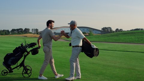 Golf players shaking hands on grass field. Two friends meeting play on weekend. Joyful golfers team carry putters clubs on golfing competition game. Leisure outdoors activity summer sport concept. Video stock