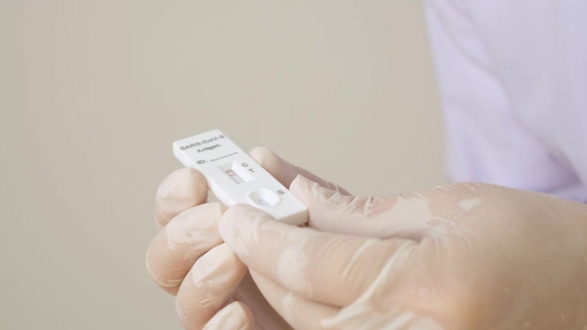 Doctor or nurse wearing rubber gloves is holding a positive Antigen Rapid Test kit (ATK) with two red lines. During a swab test for COVID-19 | Shutterstock HD Video #1091659037