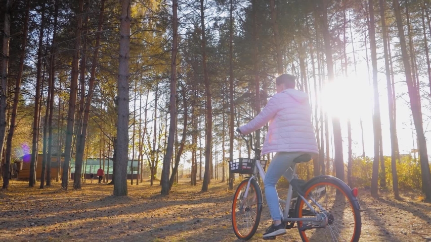 Slow motion: young woman riding bicycle in autumn city park, coniferous forest: steadicam shot. Sunset sun shines through trees - sun lens flares. Sport, freedom and active healthy lifestyle concept | Shutterstock HD Video #1091662379