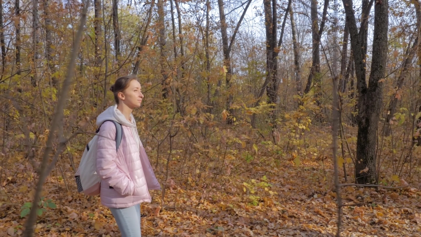 Side view of young woman in pink quilted jacket with backpack walking in autumn park, forest - wide angle steadicam shot. Active outdoor lifestyle, leisure time, freedom and adventure concept | Shutterstock HD Video #1091663389