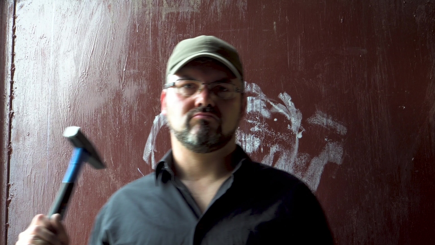 An adult man holds a hammer in his hand. A baseball cap is on his head. On the face is a beard and glasses. Worker. A dangerous person. Criminal. Preparing for crime. A carpenter. Work in the workshop