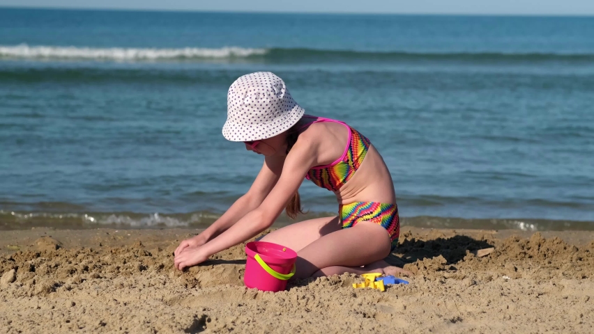 Cute child girl playing with sand on beach of ocean or sea. Childhood and summer vacation concept. Smiling kid having fun outdoors on family holidays. Kid building sand castle. Trip to warm countries | Shutterstock HD Video #1091668101
