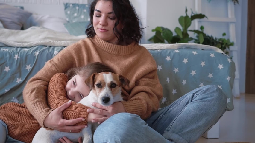 Young mother and daughter in sweaters sit on the floor on a rug with a dog jack russell and communicate cutely and play, home pastime. Pet love concept | Shutterstock HD Video #1091668221