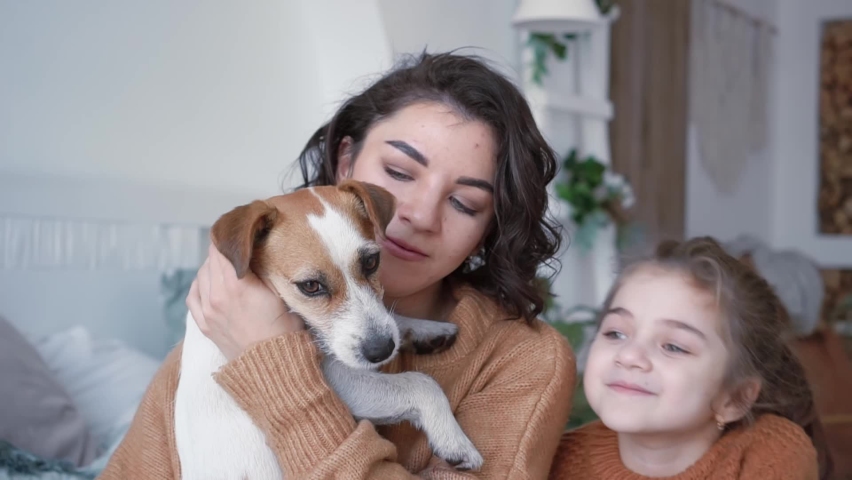 Young mother daughter in knitted sweaters are sitting on a bed in a cozy bedroom with their small dog jack russell and having a cute time with him playing with a dog. Animal love concept | Shutterstock HD Video #1091668229