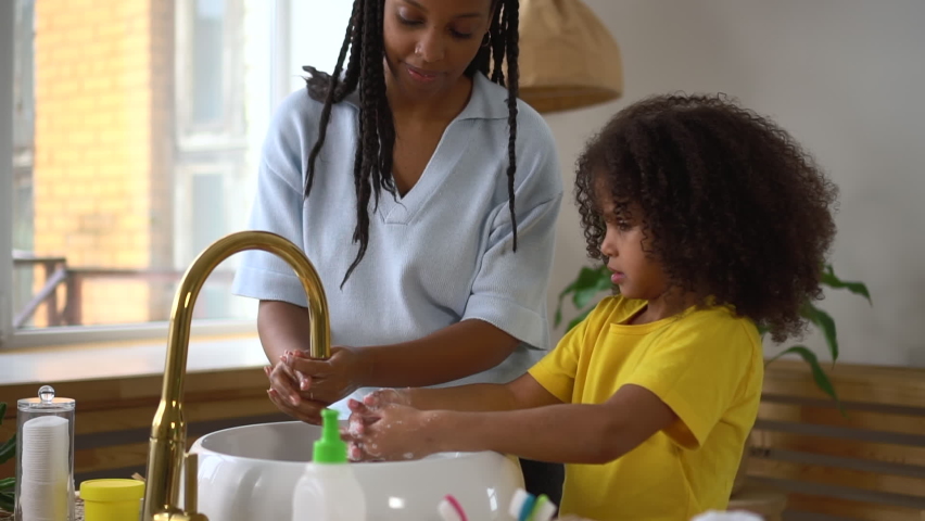 Close view of young mother, daughter are washing hands and standing in home bathroom . American african woman, girl clean hands and spread foam, talk and stand by sink in light interior. Two