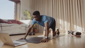 Video of a young Black African American male working out at home in his living room, doing push ups on a yoga mat. Following an online class using his laptop.