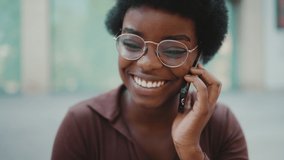 Tracking shot of attractive African dark haired girl in glasses looking happy having conversation with friend on mobile phone outdoors. Young woman using smartphone