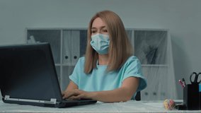 Young woman puts a medical mask on her face and works at a laptop in the office.