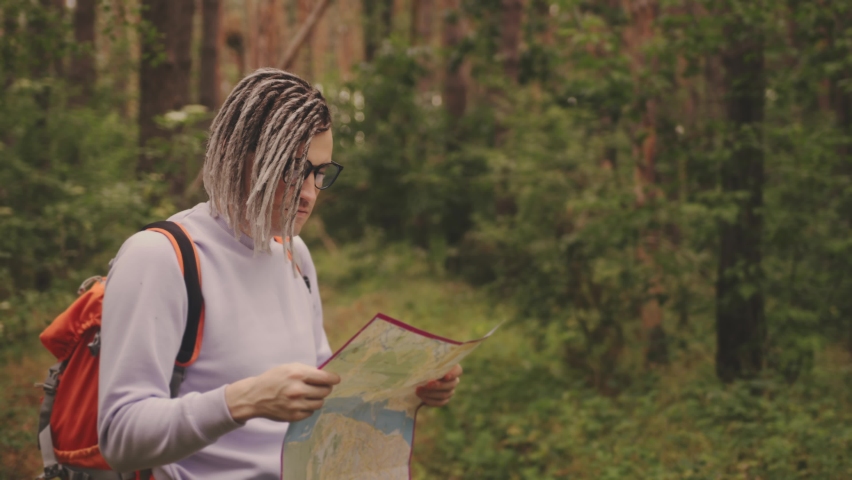 Traveling man with dreadlocks with map in woods. Male traveler with backpack in woods reading map in cloudy weather