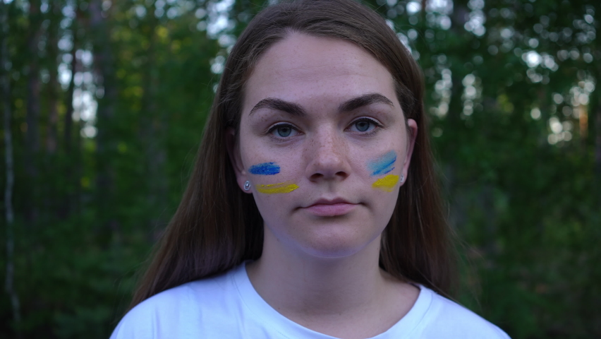 Headshot of young brunette Ukrainian woman with blue and yellow strips painted on face looking at camera. Close-up portrait of sad devastated lady posing outdoors in forest