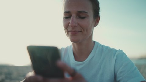 Attractive blond woman chatting with friends using smartphone outdoors. Sporty woman resting on the beach. Distant communication