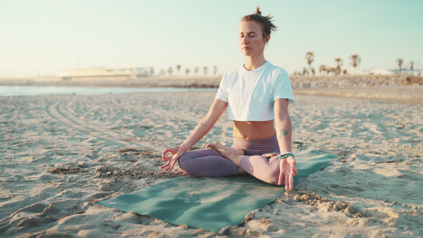 Beautiful blond yogi girl dressed in sportswear sitting in lotus pose on mat by the sea. Young tattooed woman meditating on the beach. Morning yoga outdoors | Shutterstock HD Video #1091692489
