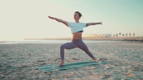Fit woman doing yoga on mat outdoors. Attractive yogi girl in sportswear standing in warrior yoga pose on the beach. Morning yoga
