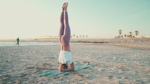 Amazing shot of strong yogi girl standing on her head doing yoga poses by the sea. Attractive woman practicing morning yoga on the beach