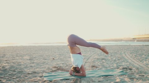 Beautiful shot of strong yogi girl doing headstand practicing yoga poses by the sea. Attractive woman keeping balance during practice yoga on the beach