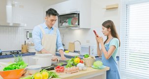 Healthy asian young couple wearing apron are cooking and woman taking videos through mobile phone in light white interior style kitchen at home