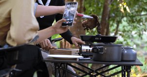 Young asian woman cooking and her friend enjoy to use smartphone record video the meal in pan, They are talk and laugh with fun together while camping in nature park