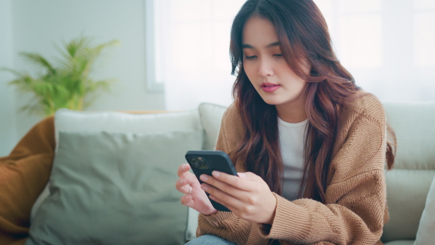 Upset young asian woman looking at mobile phone screen feels sad and disappointed. Unhappy female receive bad news while sitting on couch at home Royalty-Free Stock Footage #1091697961
