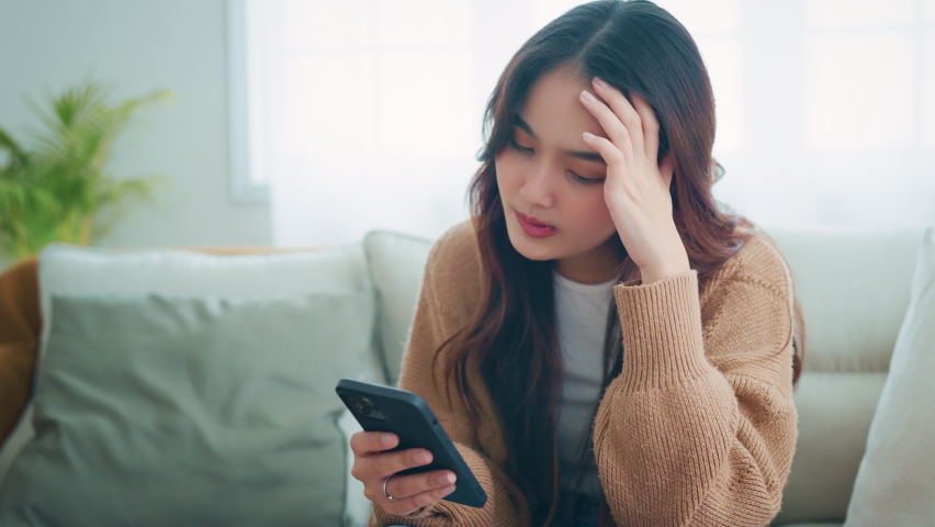 Upset young asian woman looking at mobile phone screen feels sad and disappointed. Unhappy female receive bad news while sitting on couch at home | Shutterstock HD Video #1091697961