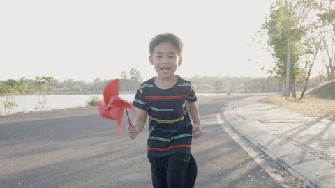 Asian child boy running and playing with pinwheels, Happiness little boy smiling in wheat field holding small wind wheel or windmill toy on hand in summer day at sunset in the park garden