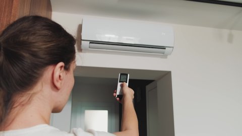 Young happy woman turns on air conditioner. Back view, casual wear t-shirt. Adult girl using remote control presses buttons, sets temperature of air cooling in house, modern technologies. White room