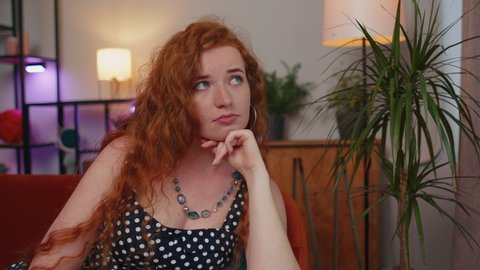 Close-up of sad young redhead woman sitting at home looks pensive thinks over life concerns or unrequited love, suffers from unfair situation. Problem, break up, depressed feeling bad annoyed, burnout