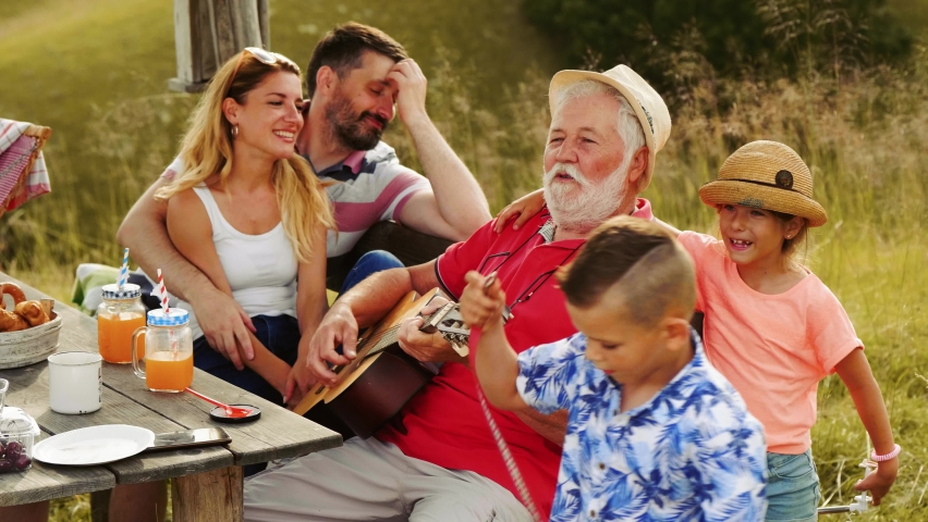 Cute little girl hugs her grandfather who plays guitar at a picnic lunch in nature. Royalty-Free Stock Footage #1091735441