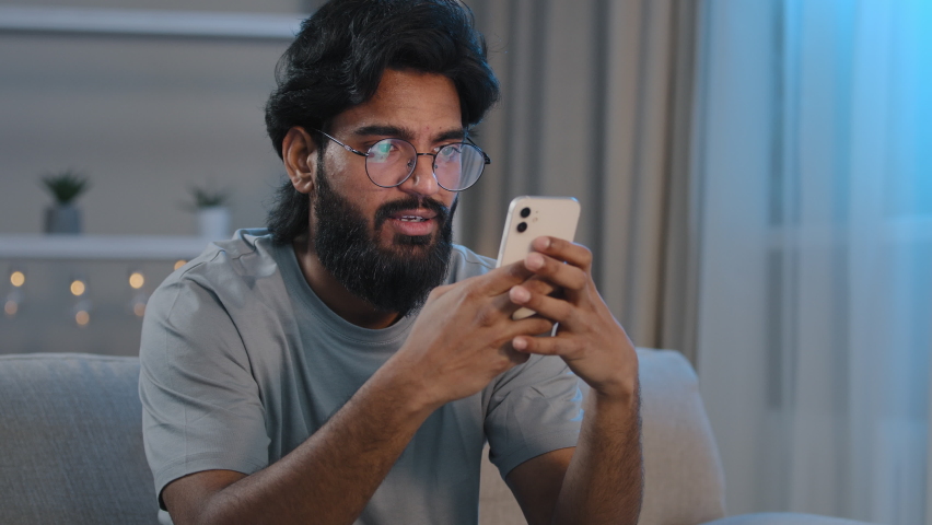 Serious busy Arabian Indian man bearded male in glasses typing browsing in smartphone texting chatting using mobile app messaging at home sitting on couch at night playing game e-commerce shopping