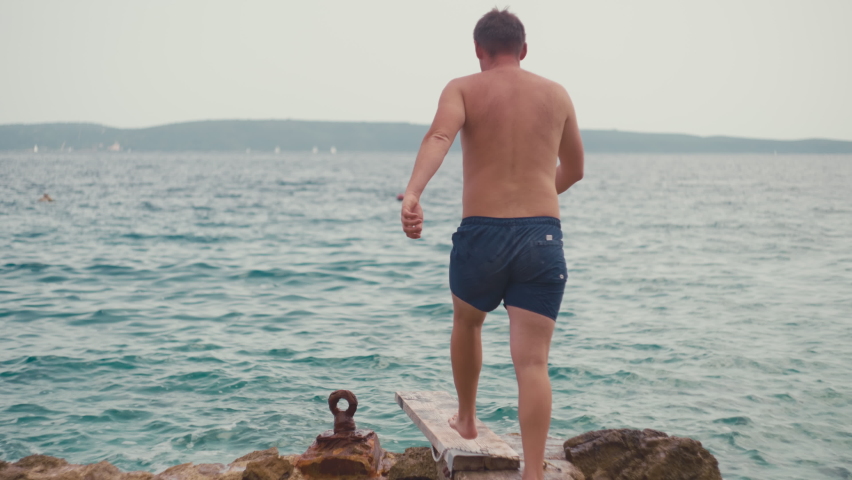 Lifestyle slow motion video of a guy jumping from a pier into the sea water. Active vacation by the ocean, happy weekend.