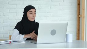 A young woman with a pleasant appearance in a black hijab communicates via video communication in a laptop, smiles during communication, gestures. Online education at the university.