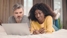 Happy middle aged man and young african american woman using laptop computer at home watching online show lying in bed together. Couple looking at gadget screen making video call chatting in bedroom - Powered by Shutterstock - Get 15% off with code: PIKWIZARD15