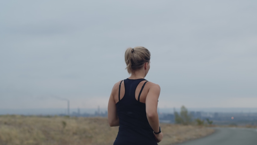 Caucasian athlete runs along side of road against the background of city and cloudy sky, rear view. A female runner trains by running on the street. Preparation for marathon | Shutterstock HD Video #1091746541