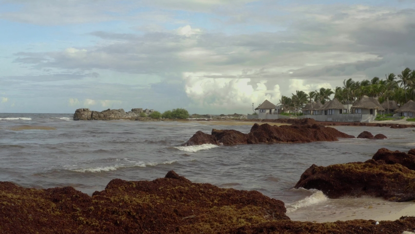 Pan shot of rocky of tropical beach in Tulum, Mexico on a cloudy day. Beautiful huts along the shoreline with waves crashing on the sandy beach.