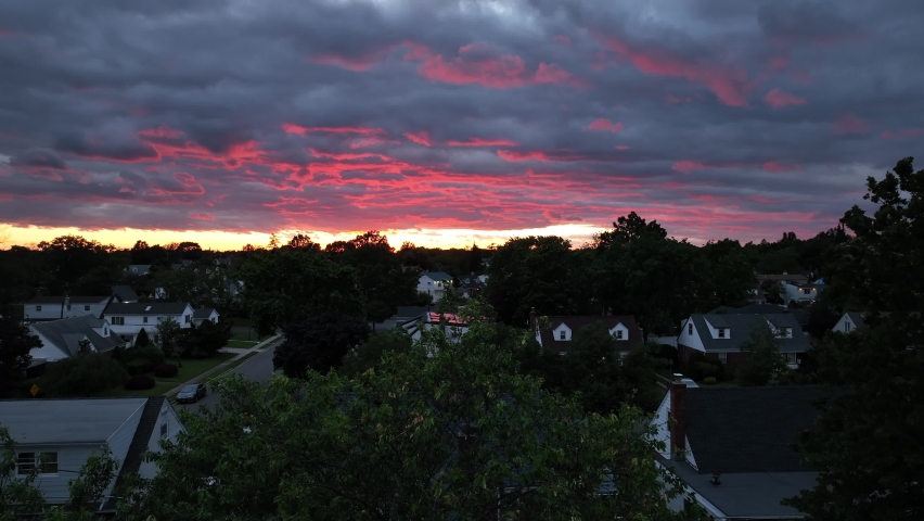 A drone view of a beautiful sunset from a suburban backyard. The neighborhood is getting dark as the camera boom down below the horizon, the golden sunset and colorful clouds down into the trees.