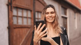 Attractive blonde curly woman with two thin braids in black clothes recording travel video blog on the old street, curl hair on finger, telling with camera and  point on something. Selective focus.