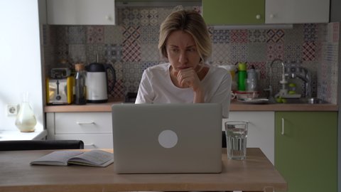 Pensive woman mature-aged job seeker sitting at kitchen table using laptop, focused middle-aged female freelancer doing remote work, working remotely at home office. Job searching after age 40