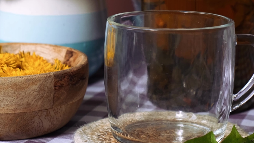 Dandelion flower healthy tea in glass teapot and glass cup on table. Delicious herbal tea pouring from fresh dandelion flowers at home at summer day near garden. Green clearing Hot dandelion tea is