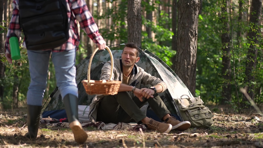 Positive handsome Caucasian man sitting at camp tent greeting young woman returning from forest with mushrooms in wicker basket. Happy couple enjoying weekend leisure in wood on spring autumn day | Shutterstock HD Video #1091769261